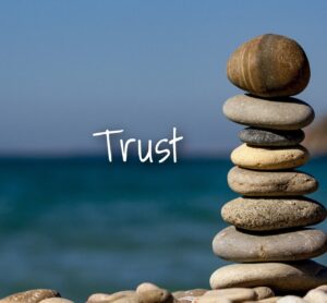 8 small flat pebbles stacked into a tower with a water scene of the ocean or river or lake in the background with the word trust on the horizon